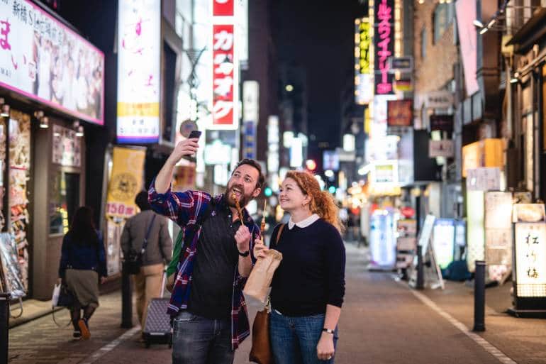 Caucasian Couple Smiling To The Cellphone Camera While Holding Street Food At A Street Outside In Japan